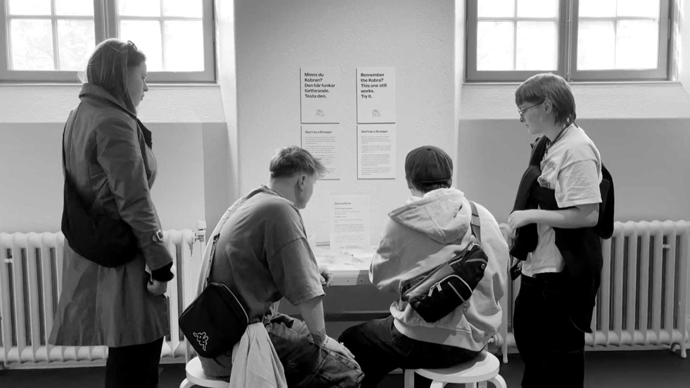Four visitors gathered round the Röhsska table, two sitting on the stools, under the yellow posters.