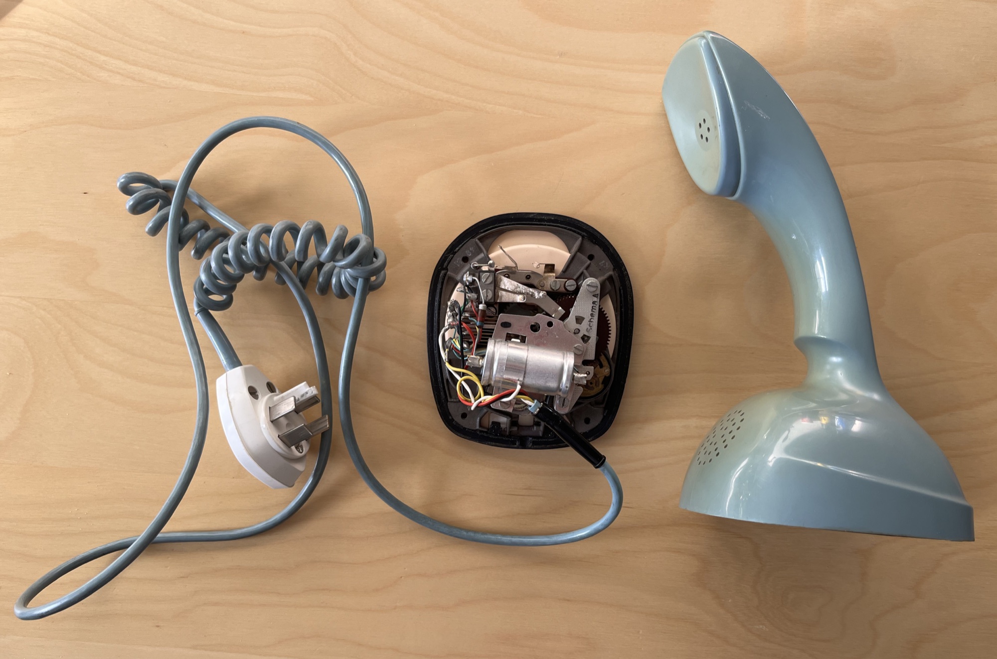A pale blue Ericofon beside its opened base, the dial mechanism showing, and its coiled cable, laid out for repair.