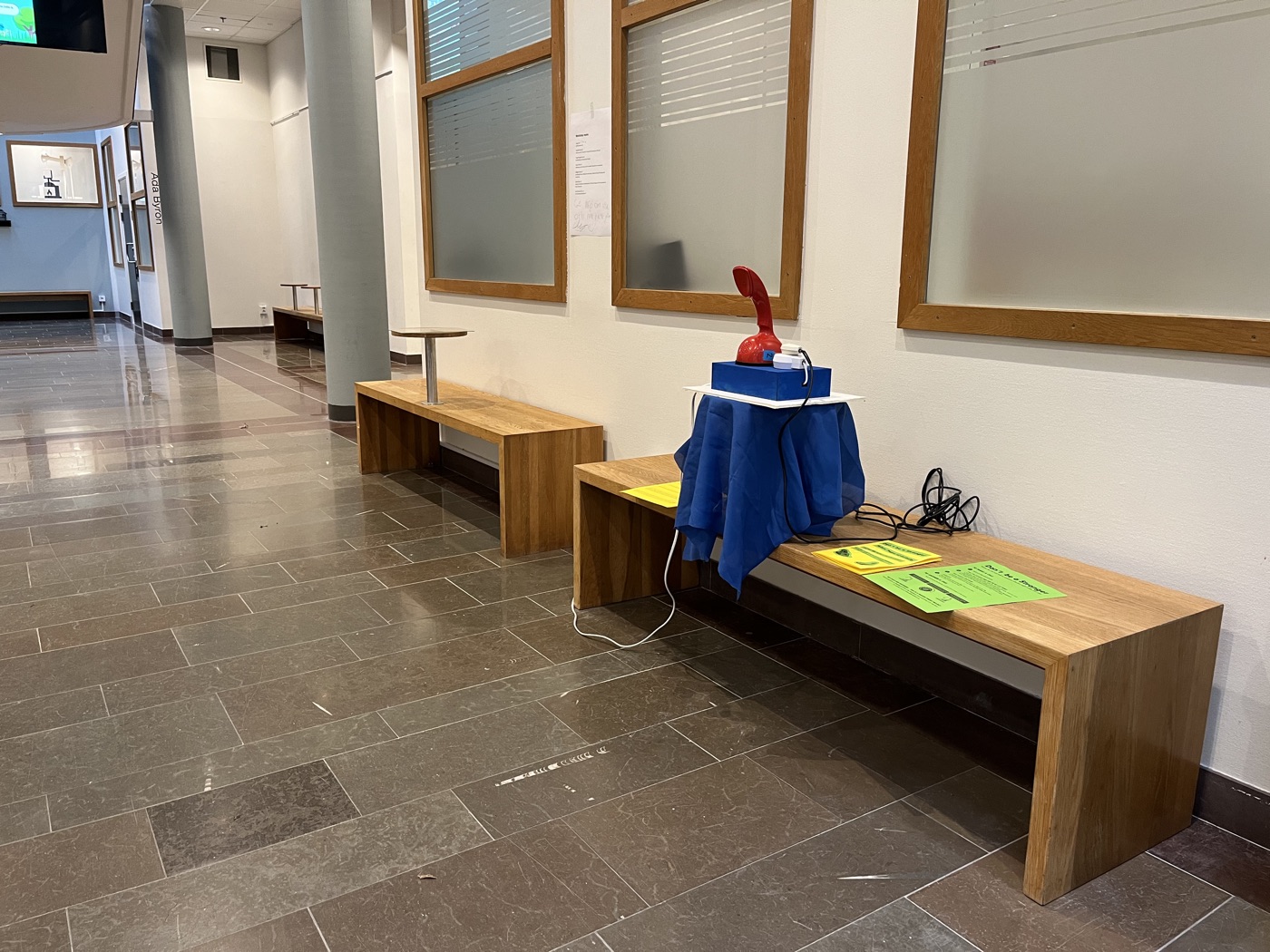A red Ericofon on a blue-draped stand at the end of a wooden bench in a long, quiet corridor.