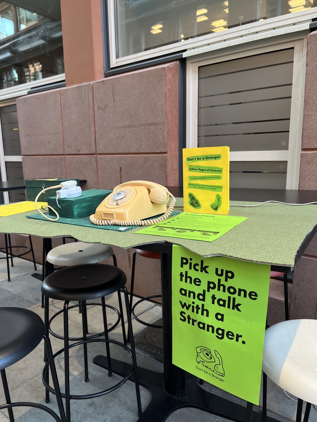 The Dialog on green felt with a large green poster hanging from the table: Pick up the phone and talk with a stranger.
