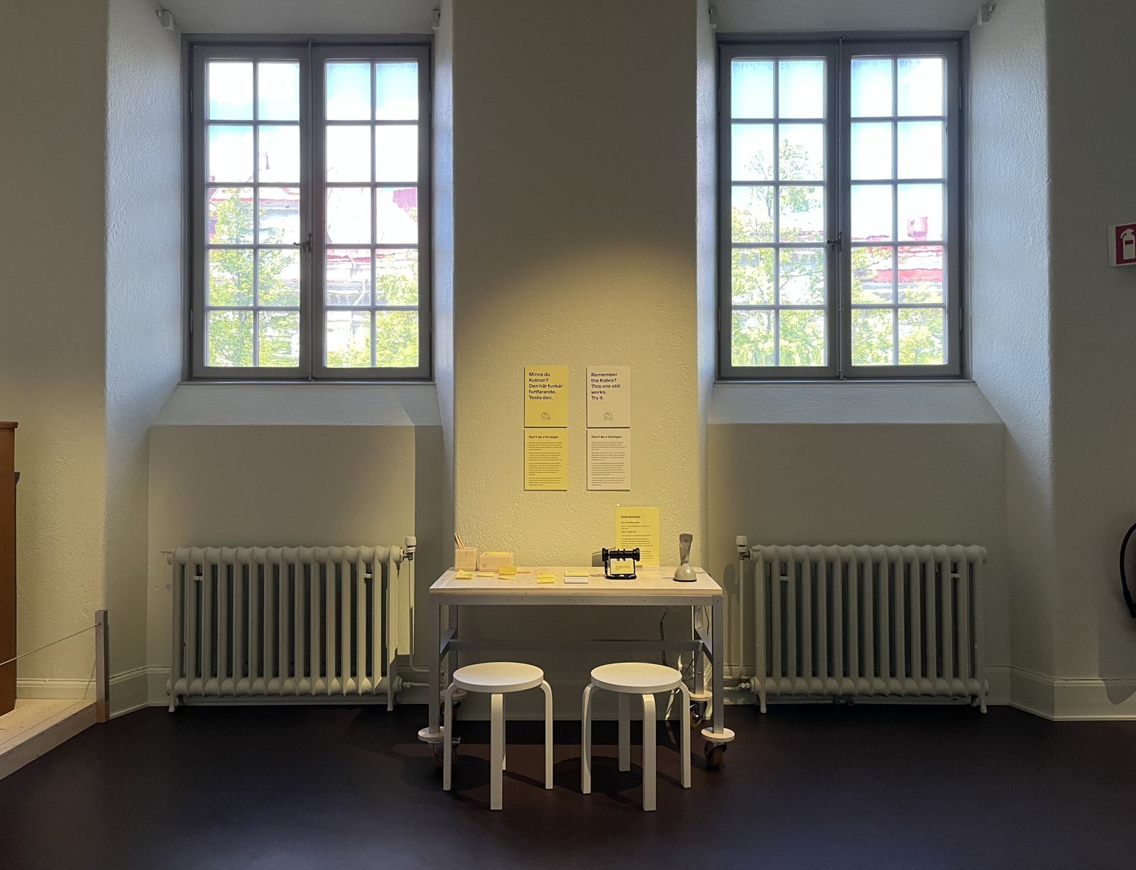 The Röhsska installation in the Innovation room: a small white table with the Ericofon, post-its and two stools, between two tall windows, with yellow posters on the wall.