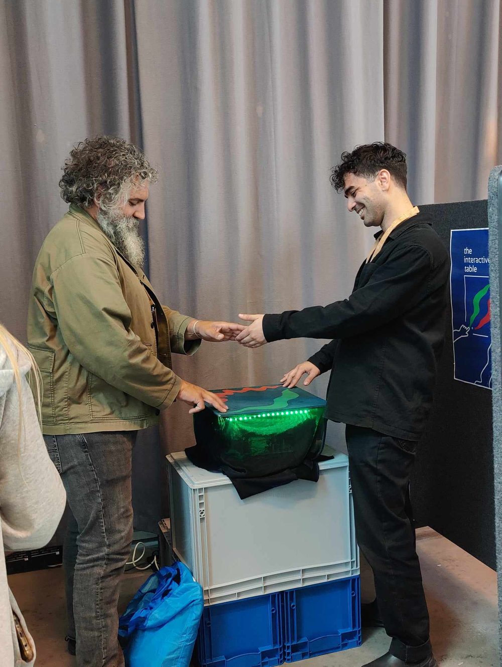 Nikos and David J. Cuartielles standing either side of the table on a crate, shaking hands across it while each touches a point, the table's green light glowing.