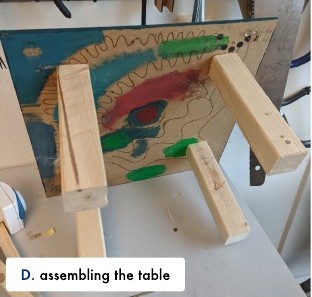D: the painted tabletop upside down on wooden legs during assembly.