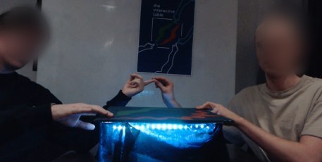 Two students touching knuckles above the table, the light underneath blue.