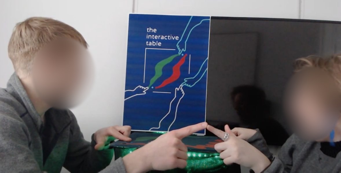 Two students facing each other across the table, each touching a point with one hand while their other fingertips meet in the middle, in front of the blue 'interactive table' poster.